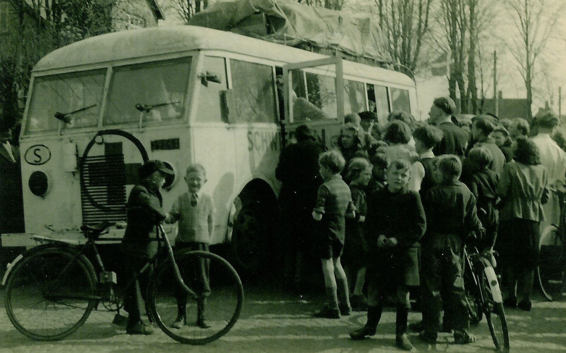 Sort hvitt bilde av gammeldags hvit buss hvor mange mennesker trenger seg sammen utenfor bussen