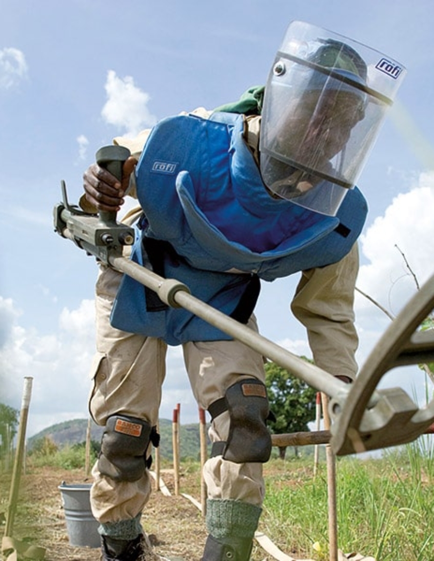 En person med sikkerhetsutstyr og metalldetektor leter i gresset.