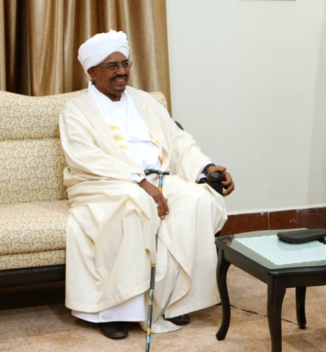 En mann i hvitt tøy og hvit turban, sitter smilende på en sofa.