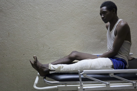 En mann i blå singlet sees fra siden. han sitter på enden av en sykehusseng med det ene benet utstrakt. benet er gipset fra foten til hoften.