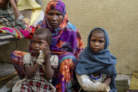 En mor i en fargerik hijab sitter med sine to små døtre ved siden av seg. Begge jentene har gips på armene. De kikker alvorlig inn i kameraet.