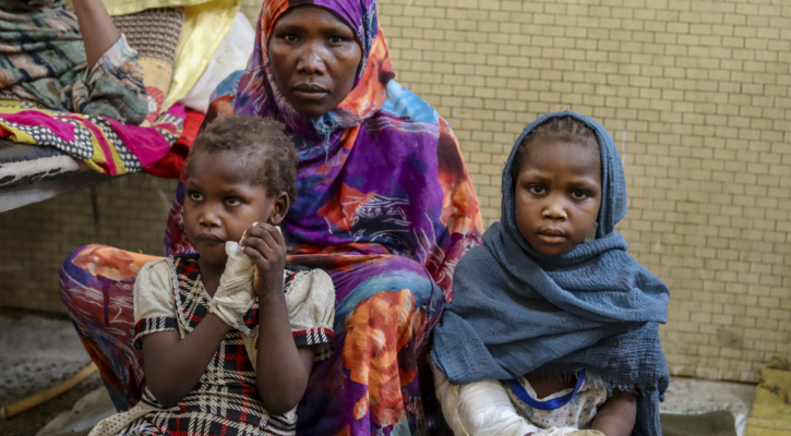 En mor i en fargerik hijab sitter med sine to små døtre ved siden av seg. Begge jentene har gips på armene. De kikker alvorlig inn i kameraet.