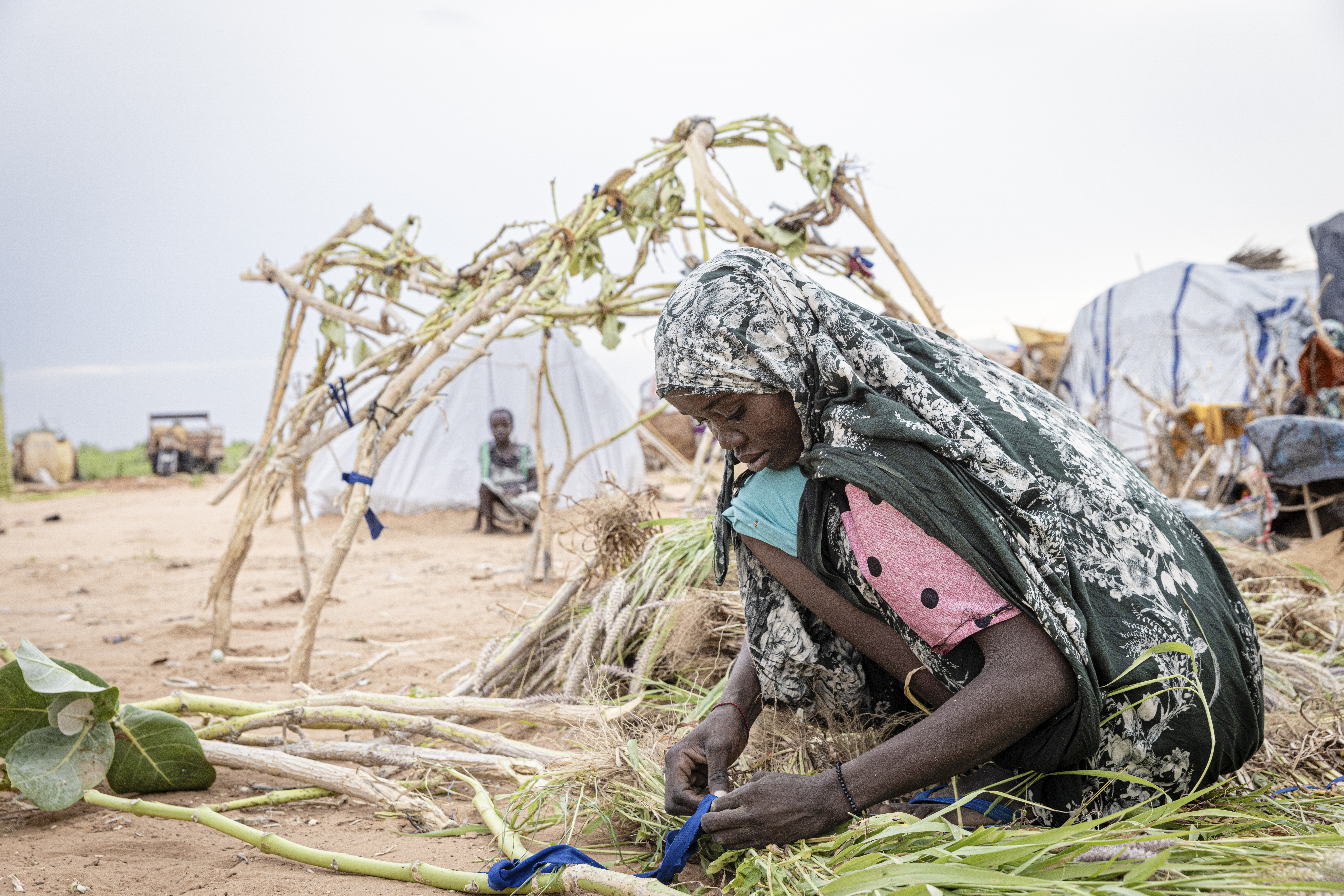 En ung sudanesisk jente med rosa t-skjorte og et sjal på hodet sitter på bakken og samler bunter med pinner og halm. I bakgrunnen er det flere telt.