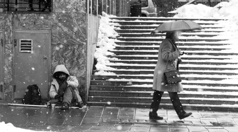 Sort/hvitt bilde av et menneske som sitter lent mot en vegg mens et annet menneske med paraply går forbi