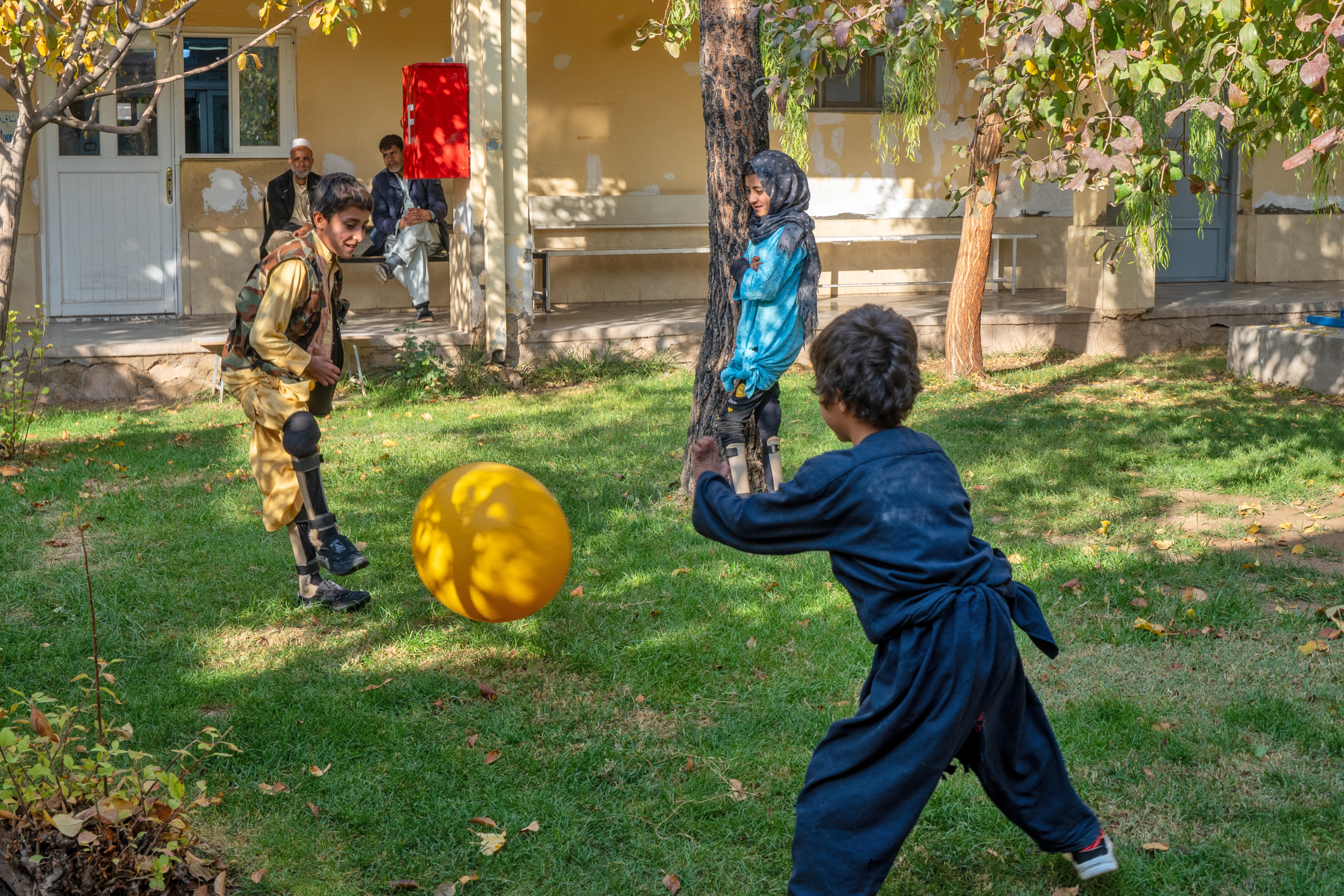 To gutter og ei jente leker med en ball i en hage foran et gult murhus. To av dem har synlige skinner eller proteser på bena.