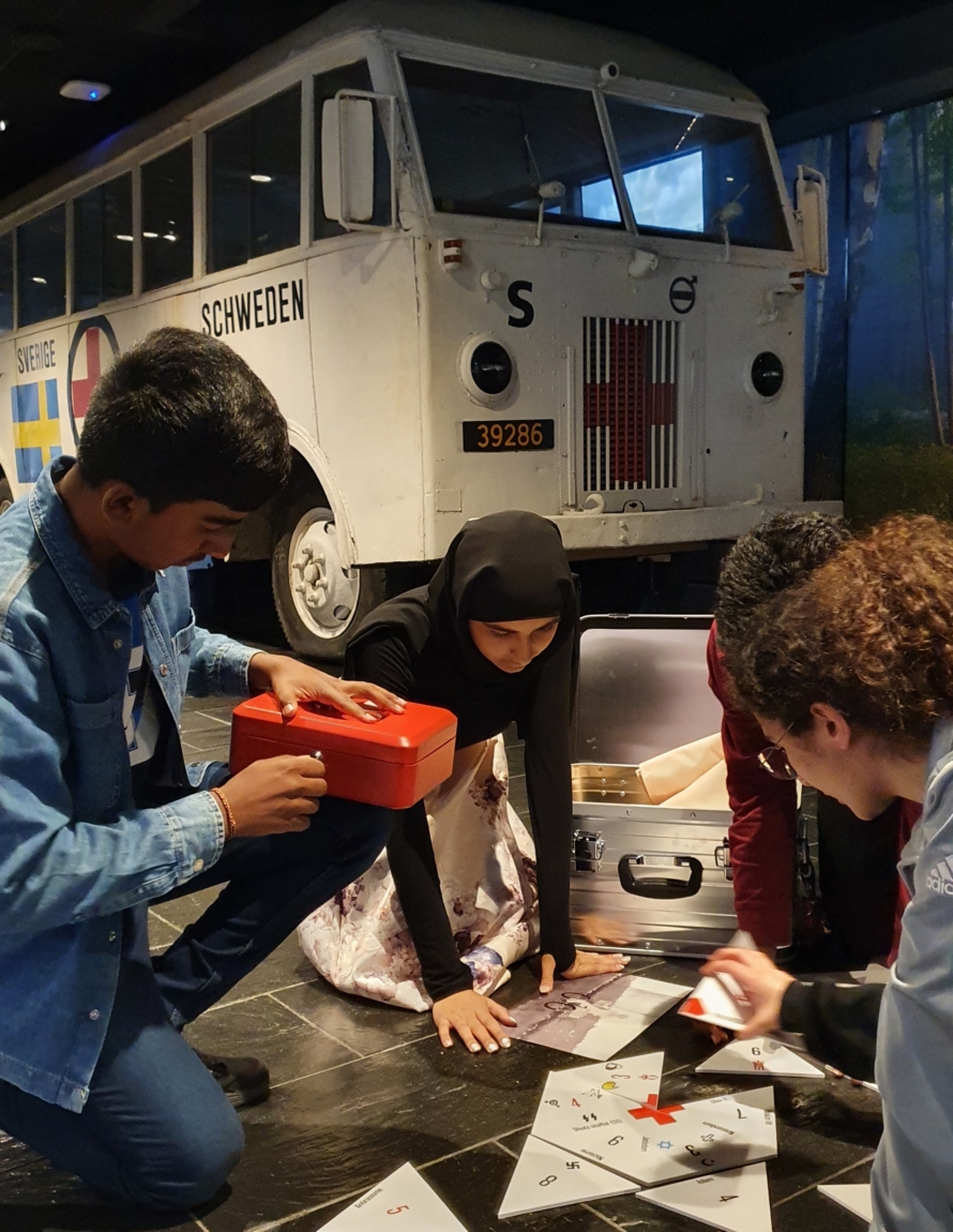 Fire ungdommer sitter på gulvet og jobber med noe som kan ligne et puslespill og det ligner åpne hengelåser på gulvet ved siden av. I bakgrunnen en hvit buss med Røde Kors-logo.