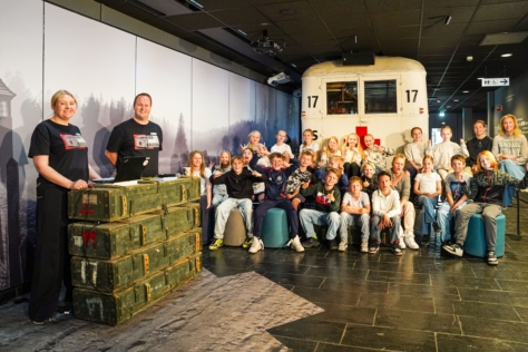 To lærere til venstre i bilde. En skoleklasse sitter og vinker til kameraet. I bakgrunnen en hvitmalt buss med Røde Kors-logo.