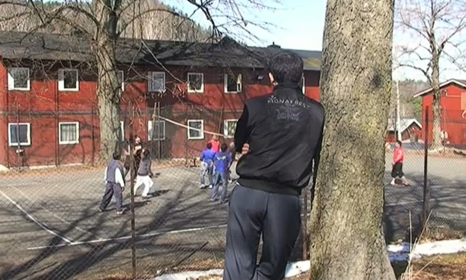 En gutt med ryggen til kameraet står på avstand og ser på en gjeng gutter spille fotball på en grusbane.