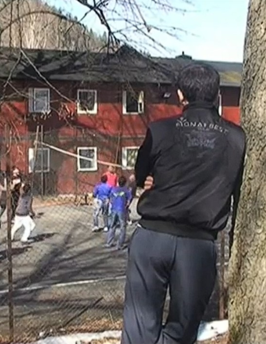 En gutt med ryggen til kameraet står på avstand og ser på en gjeng gutter spille fotball på en grusbane.