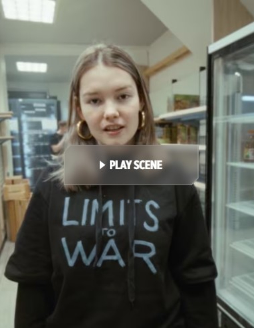 En jente står inne i et lokale som ligner en butikk, men med tomme hyller. Hun har på en t-skjorte der det står Limits to war