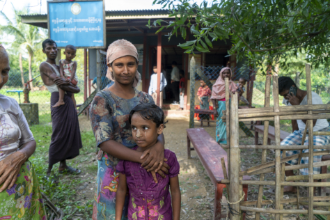 En mor og datter poserer for kameraet. Moren holder armene rundt datteren. Bak dem sees et lite bygg. Flere mennesker står i kø foran mennesker i vester med røde kors på.