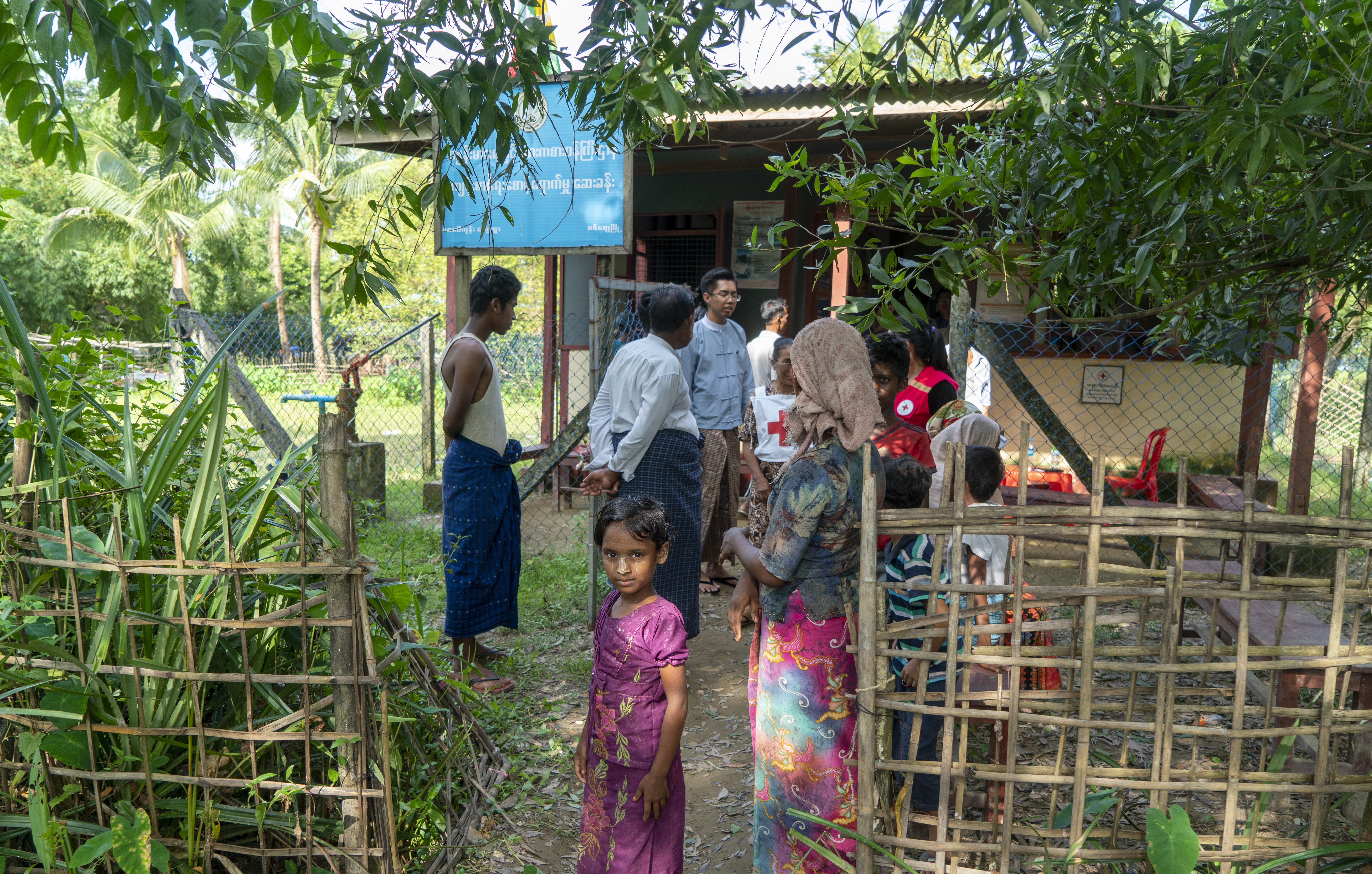 Ei lita jente i lilla kjole i frodige, grønne omgivelser. Bak henne sees et lite bygg. Flere mennesker står i kø foran mennesker i vester med røde kors på.