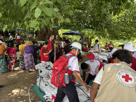 Hjelpearbeidere med Myanmar Røde Kors på ryggen organiserer mange store pakker med nødhjelp merket med røde kors. I bakgrunnen står mange mennesker som venter på hjelp.