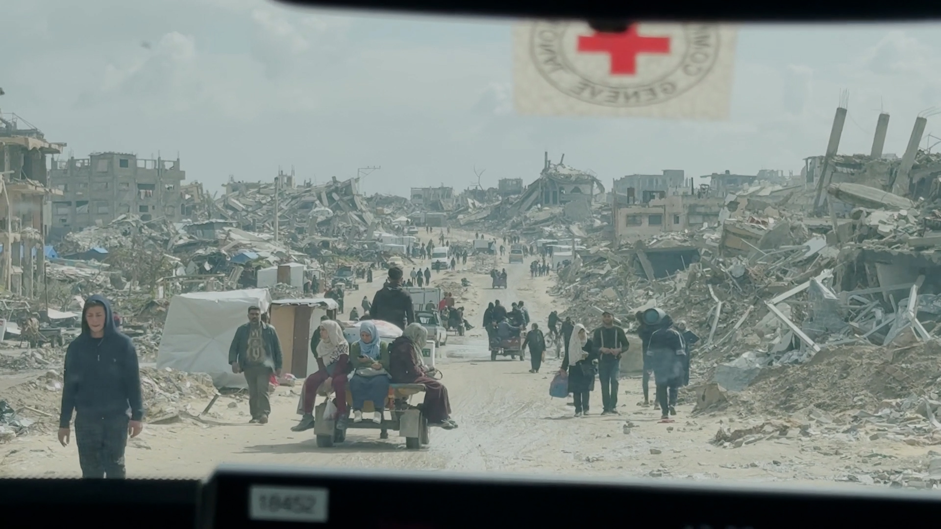 Et bilde tatt fra innsiden av en bil. På bilruten ser man et Røde Kors-merke. Bildet er fra Gaza og viser en lang grusvei med ødelagte bygninger på hver side. Veien er full av mennesker på flukt.